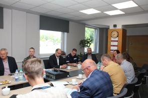 Kilkanaście osób uczestniczy w spotkaniu przy dużym stole konferencyjnym. Spotkanie odbywa się w jasnej sali z dużymi oknami.