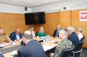 Kilkanaście osób uczestniczy w spotkaniu przy dużym stole konferencyjnym. W tle znajdują się drewniane panele ścienne, duży ekran oraz godło Polski.