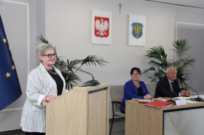Beata Macura Członkini Zarządu stoi przy mównicy i przemawia do uczestników spotkania. Przy stole konferencyjnym siedzą dwie osoby, a na blacie znajdują się dokumenty, notatniki, mikrofony oraz czerwony segregator. 