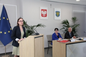 Wioletta Beyga refleksolog stoi przy mównicy. Za nią, przy stole konferencyjnym, siedzą dwie osoby, a na blacie znajdują się dokumenty. W tle widoczna jest flaga Unii Europejskiej.