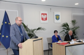 Zenon Woźniak Kierownik Biura Bezpieczeństwa i Zarządzania Kryzysowego stoi przy mównicy i przemawia. Przy stole konferencyjnym siedzą dwie osoby, a na blacie znajdują się dokumenty, notatki, mikrofony oraz czerwony segregator. W tle widoczna jest flaga Unii Europejskiej.