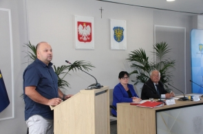 Marcin Lis fizjoterapeuta stoi przy mównicy i przemawia. Za nim, przy stole konferencyjnym, siedzą dwie osoby. W sali widoczne są mikrofony, dokumenty oraz tabliczki z nazwiskami.