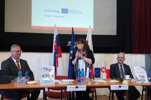 Przy stole konferencyjnym siedzą uczestnicy spotkania. Jedna z osób przemawia do mikrofonu. Na stole znajdują się materiały informacyjne, a w tle widoczne są flagi.