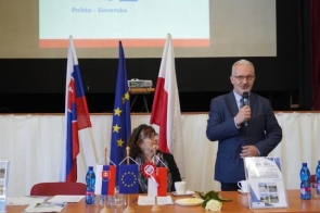 Osoba przemawiająca do mikrofonu przy stole konferencyjnym. W tle znajdują się flagi Polski, Słowacji i Unii Europejskiej oraz ekran projekcyjny z wyświetloną prezentacją.