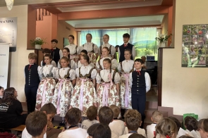 Grupa dzieci i młodzieży w strojach regionalnych stoi na scenie przed zgromadzoną publicznością. Uczestnicy ustawieni są w kilku rzędach, a w tle widoczna jest dekoracja oraz ekran z wyświetlanym krajobrazem.