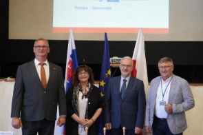 Cztery osoby pozujące do wspólnej fotografii. W tle znajdują się flagi Polski, Słowacji i Unii Europejskiej oraz ekran projekcyjny z wyświetloną prezentacją. Na pierwszym planie widoczny jest stół z materiałami informacyjnymi i flagami stołowymi.