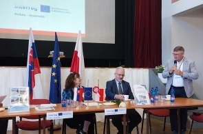 Przy stole konferencyjnym znajdują się uczestnicy spotkania. Jedna z osób stoi i przemawia do mikrofonu. Na stole ustawiono materiały informacyjne oraz flagi.