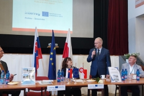 Przy stole konferencyjnym znajdują się uczestnicy spotkania. Jedna z osób stoi i przemawia do mikrofonu. Na stole ustawiono materiały informacyjne oraz flagi.