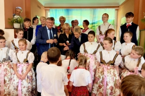Na scenie zgromadzone są dzieci i osoby dorosłe. Część uczestników ubrana jest w stroje regionalne. W centralnej części zdjęcia widoczne są osoby wręczające upominki dzieciom.