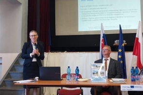 Kobieta stojąca przy stole konferencyjnym i trzymająca mikrofon. Obok siedzą uczestnicy spotkania. W tle znajdują się flagi ekran z prezentacją.