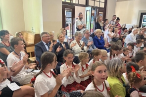 Na widowni siedzą dzieci i osoby dorosłe. Uczestnicy obserwują występ i biją brawo. W sali zgromadzona jest publiczność zajmująca miejsca siedzące oraz stojąca w tylnej części pomieszczenia.