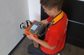 Młody uczestnik zajęć technicznych siedzi na podłodze i pracuje przy urządzeniu elektronicznym. Trzyma narzędzie w dłoni, wykonując czynności związane z montażem lub nauką elektroniki.