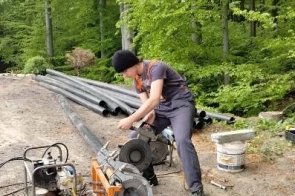 Młody mężczyzna wykonuje prace przy instalacji technicznej na zewnątrz budynku. Korzysta z narzędzi i elementów montażowych, a w tle widoczne są drzewa i inne sprzęty montażowe.