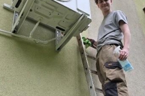 Młody mężczyzna stojący na drabinie przy klimatyzatorze zamocowanym na ścianie budynku. Ubrany w odzież roboczą.
