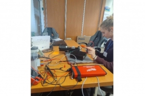 Stanowisko pracy elektronika z rozłożonymi narzędziami, przewodami i urządzeniami pomiarowymi. Przy stole siedzi osoba wykonująca prace związane z montażem lub testowaniem układów elektronicznych.