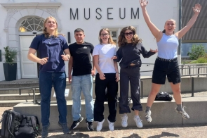 Pięć osób uchwycono podczas skoku przed budynkiem muzeum. W tle widoczna jest jasna elewacja z dużym napisem „MUSEUM”, schody oraz elementy małej architektury.