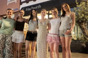 Sześć osób pozuje do zdjęcia na brukowanym placu przed budynkiem. W tle widoczne są witryny lokali usługowych oraz roślinność w dużych donicach. Zdjęcie wykonano wieczorem przy sztucznym oświetleniu.