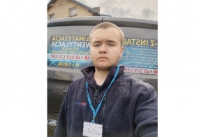 Uczestnik projektu edukacyjnego wykonuje zdjęcie typu selfie w przestrzeni publicznej. Na szyi ma identyfikator, a w tle widoczne są kolorowe plakaty informacyjne i elementy miejskiej infrastruktury.