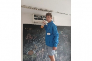 Młody mężczyzna w ręku trzyma spryskiwacz, stoi przy tablicy, nad którą wisi klimatyzator.  Ma na sobie identyfikator.