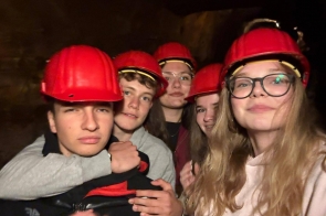 Na zdjęciu widoczna jest grupa młodych osób ubranych w czerwone kaski ochronne, pozujących do wspólnego zdjęcia we wnętrzu o ciemnych ścianach. Uczestnicy stoją blisko siebie i patrzą w obiektyw aparatu.