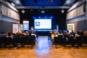 Uczestnicy siedzą na widowni sali konferencyjnej. Przed nimi znajduje się ekran prezentacyjny oraz osoba prowadząca wystąpienie.