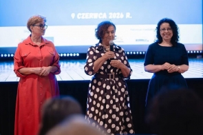 Na scenie stoją trzy kobiety. Jedna z nich trzyma mikrofon i przemawia do zgromadzonej publiczności. W tle widoczny jest ekran projekcyjny z wyświetloną prezentacją.