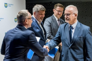 Na zdjęciu widać czterech mężczyzn. Jeden z nich podaje dłoń drugiemu, a pozostali trzymają niebieskie teczki z dokumentami. W tle znajduje się ścianka z logotypamii.