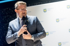 Mężczyzna w garniturze przemawia do mikrofonu na tle ścianki z logotypami Województwa Śląskiego.