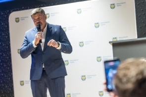 Mężczyzna w garniturze przemawia do mikrofonu na tle ścianki z logotypami Województwa Śląskiego.