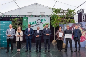 Grupa osób stoi na scenie podczas wydarzenia. Dwie osoby trzymają dyplomy i tablicę z napisem „Zielona Pracownia”. W tle widoczny jest baner wydarzenia oraz elementy scenografii.