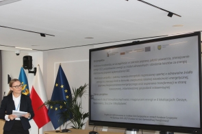 Kobieta stoi obok dużego ekranu i trzyma w dłoniach kartki. Na ekranie wyświetlana jest prezentacja dotycząca projektu związanego z odnawialnymi źródłami energii, poprawą jakości powietrza i budową instalacji fotowoltaicznych. W tle widoczne są flagi.