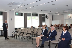 Mężczyzna stoi z przodu sali i zwraca się do siedzących uczestników. W rzędach krzeseł widoczne są kobiety i mężczyźni, część z nich trzyma kartki lub dokumenty. W tle znajdują się duże okna, głośnik i zegar na ścianie.