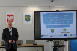 Mężczyzna stoi obok dużego ekranu, na którym wyświetlana jest prezentacja „Odnawialne źródła energii (OZE) w bilansie energetycznym budynku – w teorii i praktyce”. Na slajdzie widnieje także nazwisko prelegenta Macieja Mierzejewskiego oraz logotypy partnerów. Na ścianie w tle znajdują się godło Polski, herb Powiatu Cieszyńskiego i krzyż.