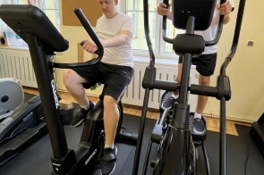 Dwóch młodych mężczyzn ćwiczy na sprzęcie treningowym w jasnej sali. Jeden siedzi na rowerku stacjonarnym, a drugi korzysta z orbitreka. W tle widoczne są duże okna oraz fragmenty pozostałego wyposażenia sali.