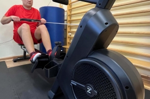 Młody mężczyzna w czerwonym stroju sportowym ćwiczy na wioślarzu treningowym w jasnej sali. Siedzi na siedzisku, trzyma uchwyt urządzenia i opiera stopy na podnóżkach. W tle widoczne są drabinki gimnastyczne oraz elementy wyposażenia sali.