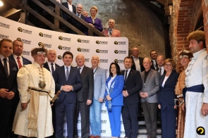 Grupa kobiet i mężczyzn stoi przed ścianką z powtarzającym się napisem „Powiat Częstochowski”. Większość osób ma na sobie garnitury lub eleganckie stroje, a po bokach stoją mężczyźni w historycznych strojach z nakryciami głowy. Część osób znajduje się wyżej, na drewnianych schodach.