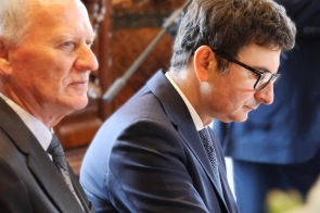 Dwóch mężczyzn w garniturach siedzi obok siebie. Jeden z nich ma siwe włosy i patrzy lekko w bok, a drugi ma ciemne włosy, okulary i spogląda w dół. W tle widoczne jest wnętrze pomieszczenia oraz fragment stojącej osoby.