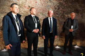 Czterech mężczyzn stoi we wnętrzu z kamienno-ceglanymi ścianami. Trzech z nich ma na sobie garnitury, a jeden ciemną kurtkę. Część osób ma identyfikatory zawieszone na szyi, a podłoga wykonana jest z metalowej kratownicy.