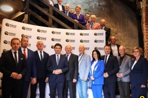 Grupa kobiet i mężczyzn stoi przed ścianką z powtarzającym się napisem „Powiat Częstochowski”. Część osób znajduje się na drewnianych schodach powyżej, pozostali stoją na dole. Większość ma na sobie garnitury lub eleganckie stroje, a w tle widoczne są kamienne ściany i drewniana konstrukcja schodów.