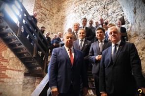 Grupa kobiet i mężczyzn stoi na drewnianych schodach we wnętrzu z kamiennymi i ceglanymi ścianami. Większość osób ma na sobie garnitury i eleganckie stroje. Na pierwszym planie stoi czterech mężczyzn, a za nimi na kolejnych stopniach widoczni są pozostali uczestnicy.