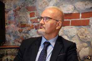 Mężczyzna w garniturze i okularach siedzi na tle kamienno-ceglanej ściany. Ma krótko ostrzyżone włosy i siwy zarost, a wzrok kieruje w bok.