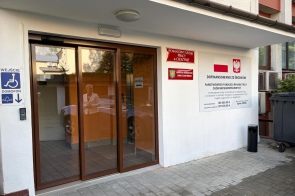 Przeszklone wejście do budynku Powiatowego Urzędu Pracy w Cieszynie. Po lewej stronie znajduje się oznaczenie wejścia dla osób z niepełnosprawnościami i domofon, a po prawej tablice z nazwą urzędu, herbem Powiatu Cieszyńskiego oraz informacją o dofinansowaniu ze środków Państwowego Funduszu Rehabilitacji Osób Niepełnosprawnych.