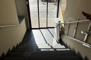 Schody prowadzą w dół do przeszklonego wejścia budynku. Po prawej stronie widoczna jest platforma przyschodowa zamontowana przy metalowej poręczy, a po obu stronach schodów znajdują się uchwyty. Przez szklane drzwi widać chodnik, ulicę i zaparkowany samochód.
