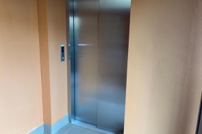 Winda z metalowymi drzwiami znajduje się w niewielkim korytarzu o jasnych ścianach. Po lewej stronie widoczny jest przycisk do wezwania windy oraz wyświetlacz pokazujący numer piętra. Podłoga wyłożona jest szarymi płytkami.