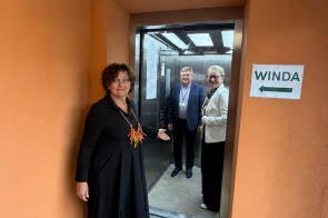 Janina Żagan Starosta Cieszyńska stoi przy otwartych drzwiach windy i gestem wskazuje jej wnętrze. W windzie stoją Jerzy Pilch Członek Zarządu Powiatu Cieszyńskiego oraz Anna Stefaniak-Bacza Dyrektor Powiatowego Urzędu Pracy w Cieszynie . Na pomarańczowej ścianie obok drzwi znajduje się kartka z napisem „WINDA” i strzałką skierowaną w lewo.