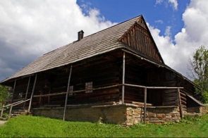 Drewniana, zabytkowa chata góralska na kamiennej podmurówce, z szerokim gankiem i balustradą z surowych żerdzi. Spadzisty, gontowy dach i ciemne, ciosane bale podkreślają tradycyjny beskidzki charakter. Wokół zielona trawa, w tle niebo z kłębami chmur.