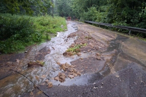 Rozmyta nawierzchnia drogi z zalegającym rumoszem i wypływającą wodą opadową. Widoczne zniszczenia i zalanie pobocza w wyniku intensywnych opadów w zalesionym terenie. W tle ustawione barierki ostrzegawcze.
