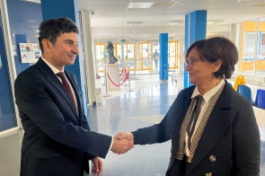 Dwójka osób w oficjalnych strojach podaje sobie dłonie w przestronnym, dobrze oświetlonym holu. Marek Wójcik Wojewoda Śląski  po lewej ma na sobie ciemny garnitur, białą koszulę i bordowy krawat. Janina Żagan Starosta Cieszyński po prawej ubrana jest w elegancką czarną marynarkę z ozdobnymi lamówkami oraz białą koszulę.