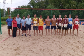 Grupa osób stoi na boisku do siatkówki plażowej. Pośrodku uśmiechnięci zawodnicy w sportowych strojach prezentują medale i dyplomy. Obok nich organizatorzy w koszulkach polo. W tle siatka i drzewa, atmosfera sportowego święta.
