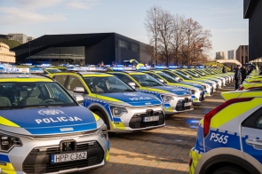 Rząd nowych policyjnych radiowozów stoi ustawiony w równych liniach na otwartej przestrzeni w słoneczny dzień. Samochody mają biało-niebieskie malowanie z żółtymi elementami odblaskowymi oraz włączone sygnały świetlne. W tle widać nowoczesne budynki, funkcjonariuszy oraz bezlistne drzewa.